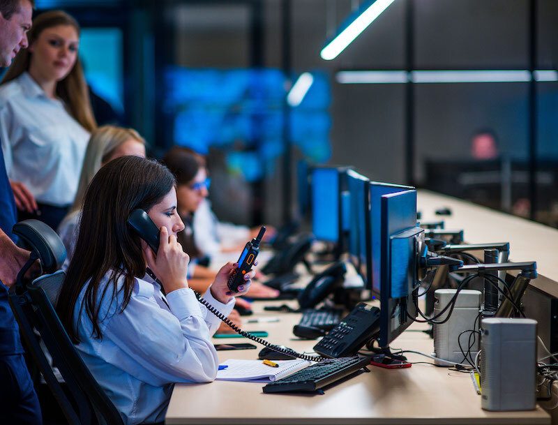 Operators at their desks in a control room, taking calls in front of a video wall.